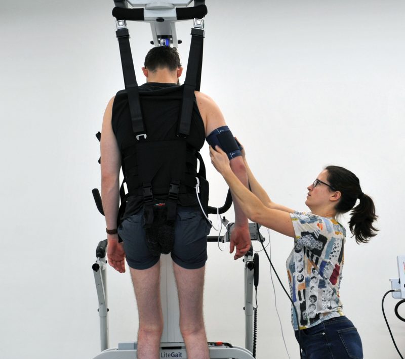 eWalk trial participant in harness on walker machine, is assisted by physiotherapist, while receiving neurostimulation.