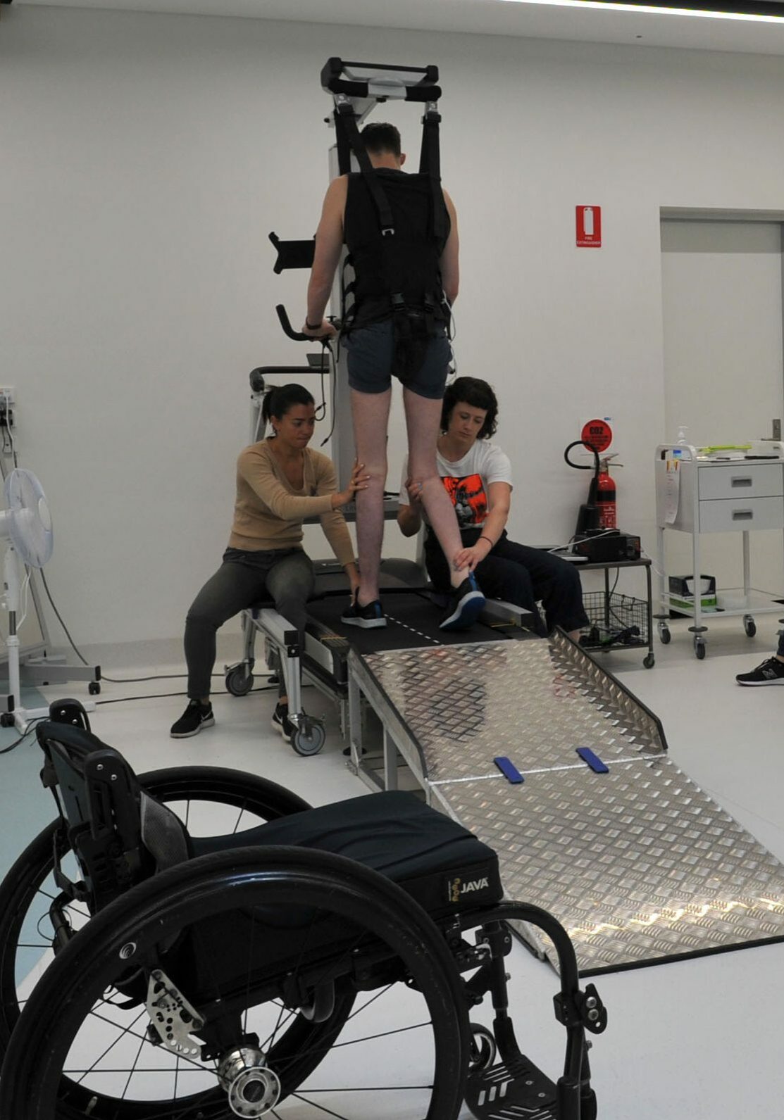 Therapy with wheelchair eWalk trial participant on treadmill with two physiotherapists and researcher, with their wheelchair in the foreground